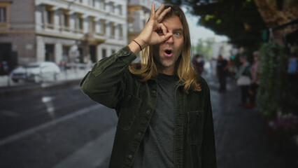 Fototapeta premium Blond man with long hair shows ok sign with his hand over eye while standing on a busy city street; approval.