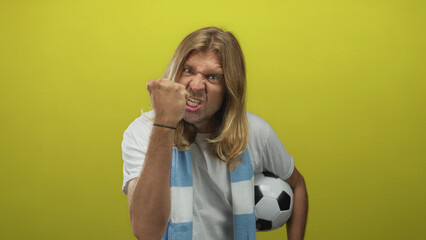 Naklejka premium Man with long blond hair holding soccer ball under arm clenches fist fiercely and snarling in studio; anger.
