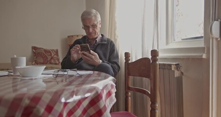 An older senior man uses his smart phone while seated comfortably at a checkered table in his home. This video captures a quiet moment of modern connection and digital interaction in everyday life. - Powered by Adobe