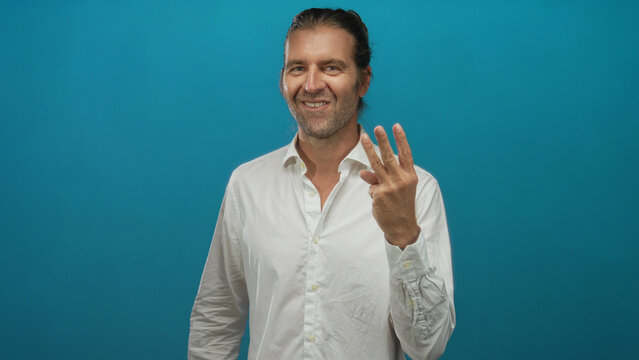Man holding three fingers up in blue studio wearing white shirt with long hair pulled back; celebration.