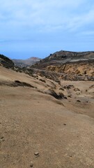 Smooth sand-colored volcanic slopes and dry scrub in Upper Teno ravine, Tenerife. Eroded cliffs and exposed soil under pale blue sky create minimalistic summer scenery.