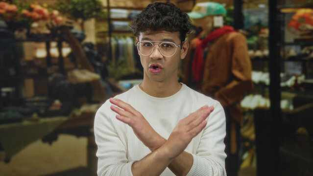 Man wearing white shirt and round glasses in clothing shop aisle crosses arms firmly in building; denial.