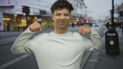 Fototapeta premium Man in white sweater wearing glasses flexes muscles and clenches fists on city street during daytime; confidence.