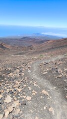Mountain trail winds over rocky volcanic slopes toward Pico Viejo, Teide National Park. Expansive view of lava fields, caldera rim, and distant Atlantic beneath the vivid blue sky.