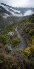 Winding mountain road curves through lush valley, trees in autumn, mist shrouds peaks under a cloudy sky