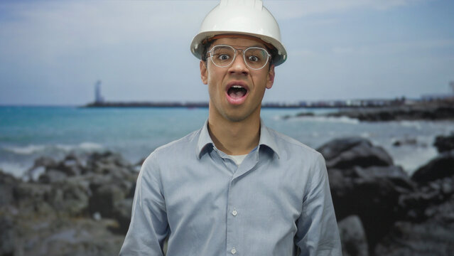 Man covering hands over mouth at building on rocky beach with ocean horizon under cloudy sky; amusement and laughter.
