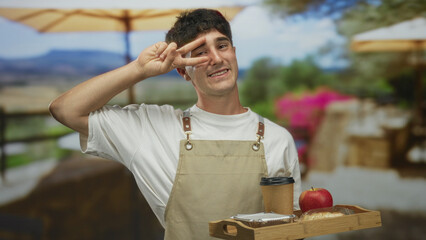 Man wearing apron holding wooden tray with coffee cup, pastry and apple on restaurant terrace, hand showing peace sign to eye; friendly service.