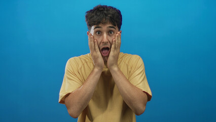 Young hispanic man pressing hands to face in studio against bright blue backdrop; surprise reaction.