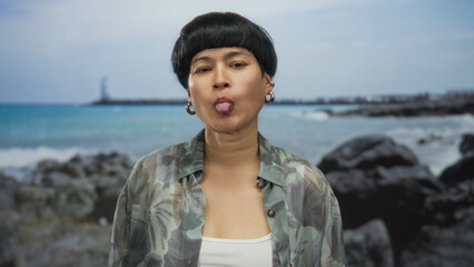 Young chinese woman outdoors at beach with sea in background playfully sticking out tongue in front of seaside rocks on a sunny day.