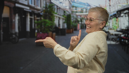 Woman points finger forward on pedestrian street while smiling and touching chest with other hand,...