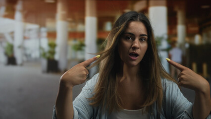 Woman points fingers to cheeks in building concourse while smiling, wearing denim shirt and tank top; confidence self expression.