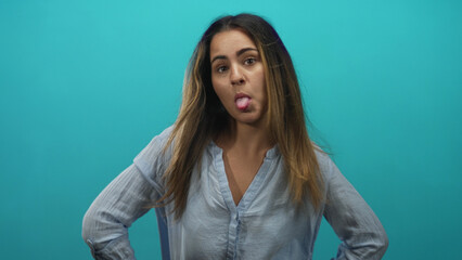 Hispanic woman sticks tongue out with hands on hips in studio against turquoise background; playful...