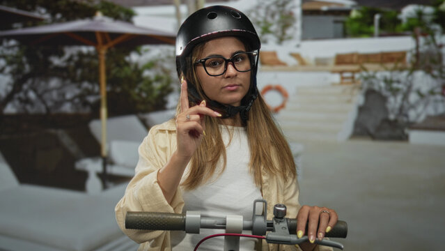 Teenage girl wearing helmet and glasses points index finger while holding scooter handlebars in studio; calm.