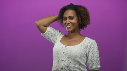 Woman wearing white knit top smiling and posing with arm bent and hand behind head in purple studio; confidence ease.