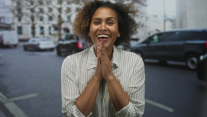 Woman smiling with clasped hands on street in city traffic facing camera and wearing striped shirt;...