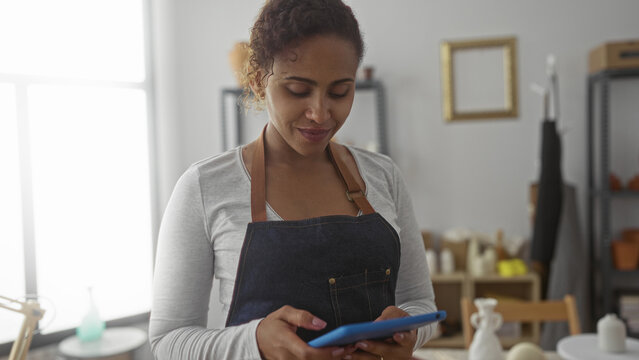 Woman holding tablet in artisan studio, smiling and tapping screen while wearing denim apron and looking down; small business creativity contentment.