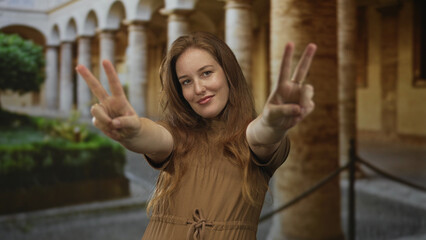 Smiling woman with red hair shows hands making peace sign on street with historic building view;...