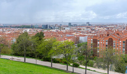 A panoramic view of the Madrid skyline from an elevated park, showing a dense expanse of terracotta rooftops and residential buildings giving way to modern high-rises in the hazy distance