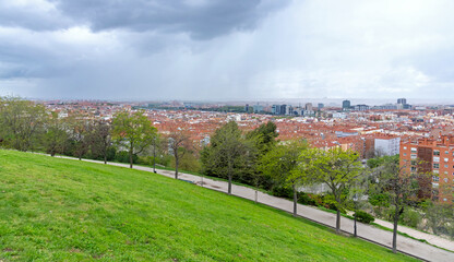 A panoramic view of the Madrid skyline from an elevated park, showing a dense expanse of terracotta rooftops and residential buildings giving way to modern high-rises in the hazy distance