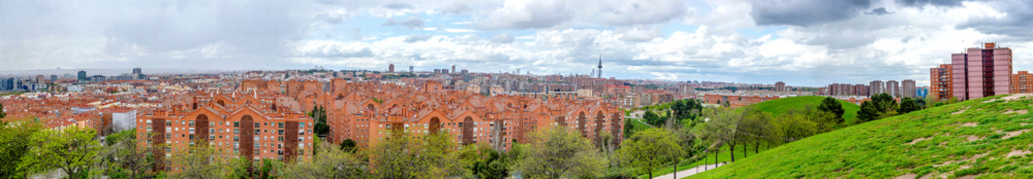 A panoramic view of the Madrid skyline from an elevated park, showing a dense expanse of terracotta rooftops and residential buildings giving way to modern high-rises in the hazy distance