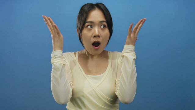 Young chinese woman wearing cream top expresses surprise with hands raised against isolated blue background wall. - Powered by Adobe