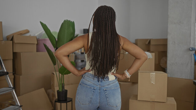 Woman in jeans and tank top with hands on hips amid stacked cardboard boxes in house; moving new home confidence.