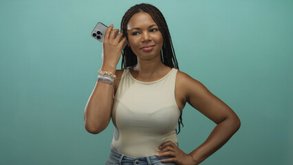 African american woman holding smartphone to ear with hand on hip and braided hair visible in teal studio; confidence connectivity technology.