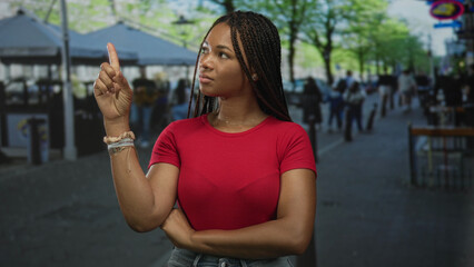 Woman points finger upward with crossed arms and visible forearm in front of outdoor cafe on a busy city street; skepticism.
