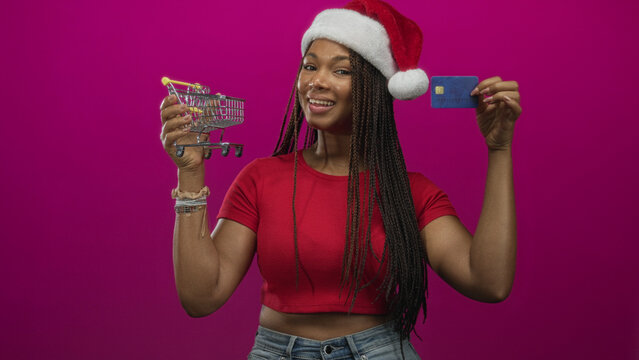 Woman wearing santa hat holding a mini shopping cart and credit card in studio with magenta backdrop; holiday shopping joy.