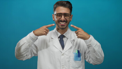 Young hispanic scientist man wearing white lab coat points fingers at teeth in blue studio; confidence.