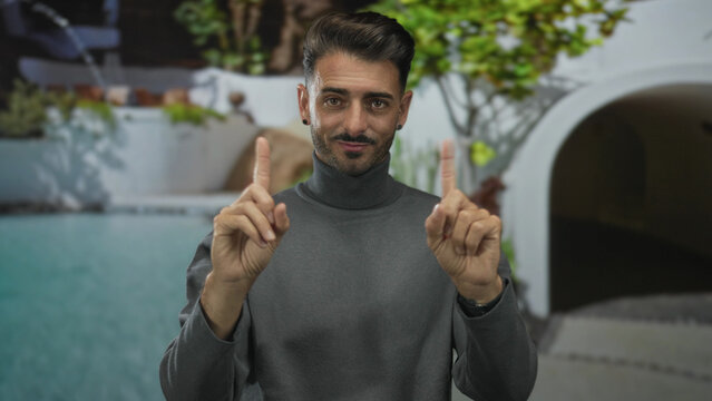 Sitting young hispanic man pointing both index fingers by turquoise pool in modern hotel building; invitation leisure.