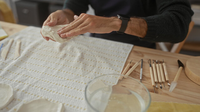 Man kneads wet clay on wooden table in studio, shaping a smooth slab with bare hands and carving tools; creativity. - Powered by Adobe