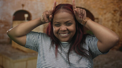 Woman with hands forming horns on head in street of old town, smiling broadly and wearing striped shirt; rock fun metal.