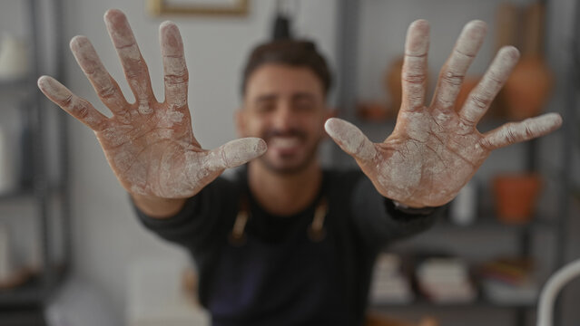 Man shows clay covered hands toward camera in artisan workshop studio wearing apron and smiling broadly; creativity passion dedication.