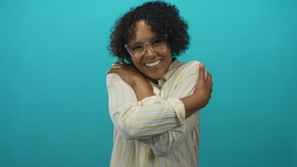 Young african american woman wearing clear eyeglasses hugging shoulders in turquoise studio; self care joy.