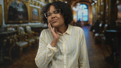 Young african american woman wearing glasses touches her cheek in museum gallery filled with paintings and chairs; distress.