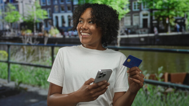 Woman holds smartphone and creditcard by a canal railing while smiling broadly on a city street; confidence. - Powered by Adobe