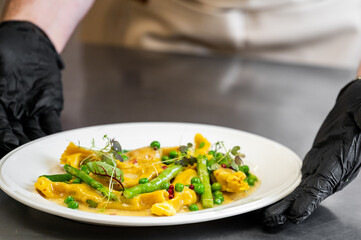 A close-up shot of a chef's hands in black gloves presenting a plate of vibrant, freshly prepared Italian pasta with asparagus, green beans, peas, and microgreens in a creamy sauce.
