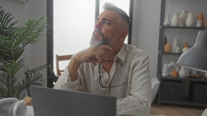 Man holding glasses with hand on chin at desk in home office, gazing upward in a pensive pose beside laptop and lamp; thoughtful reflection.