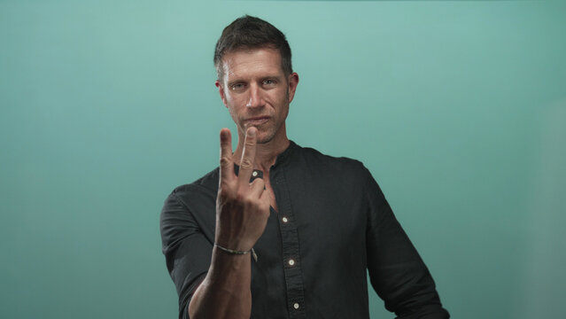 Man holding up two fingers with hands forward wearing casual black shirt and visible bracelet in studio with teal wall; playful.