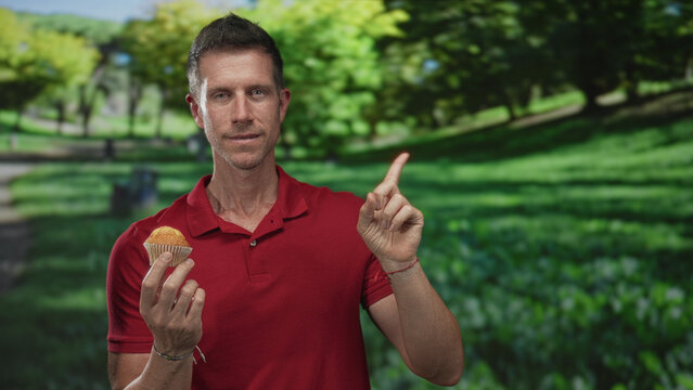 Man in red polo holding a muffin and points finger up while standing in a forest park with green trees and grass; thoughtful reflection.