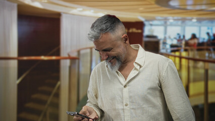 Man smiling and looking down while texting on a smartphone, hands visible in a hotel building atrium with curved railing and staircase; quiet contentment.