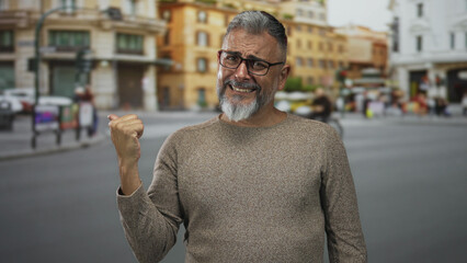 Man points thumb over shoulder on street with clenched hand, glasses and gray beard, wearing sweater in front of city buildings; skepticism.