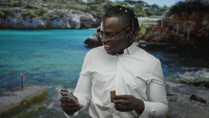 Young african american man smiles while holding smartphone and chocolate bar and checking screen in studio setting; joy casual leisure.