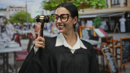 Hispanic woman judge holding gavel smiling playfully on restaurant terrace wearing uniform with...