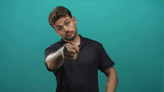 Young hispanic man pointing index finger toward camera in teal studio, visible forearm and raised hand; warning.