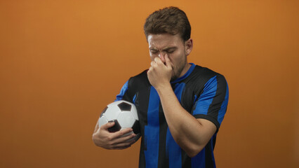 Hispanic young man pinches nose while holding a soccer ball against an orange studio wall; disgust odor reaction.