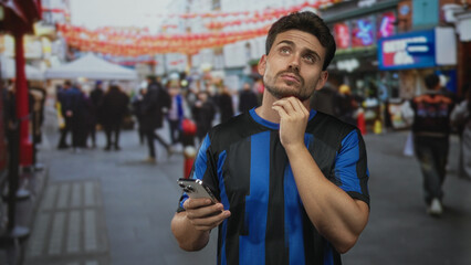 Young hispanic man holding smartphone, hand on chin while checking screen in a crowded city street...