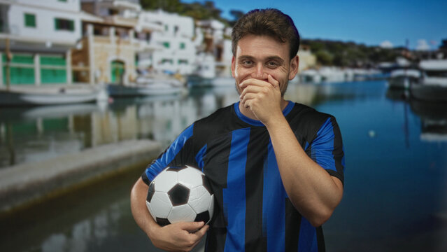 Man holding soccer ball with hand covering mouth, young hispanic wearing blue striped jersey, standing by boats at port; playful amusement team spirit. - Powered by Adobe
