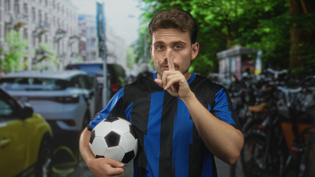 Young hispanic man holding soccer ball, finger to lips for silence on busy city street; secret quiet.